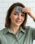Woman applying a blackhead removal pad to her forehead against a white tiled wall.